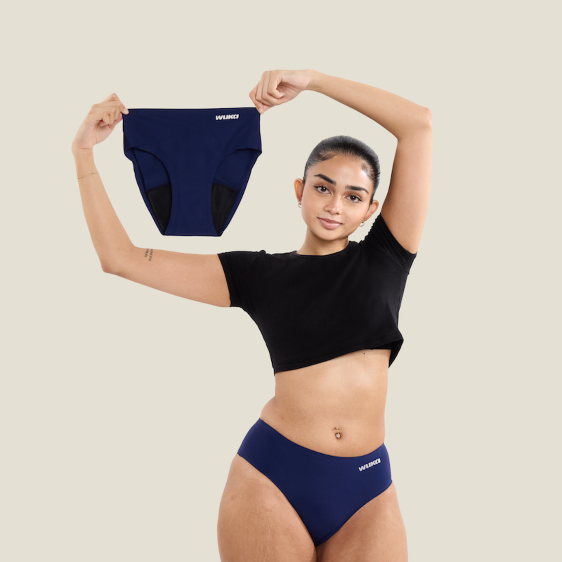 A confident teen girl in a black crop top and navy WUKA period pants holds up a matching pair, proudly showcasing reusable, sustainable period protection.